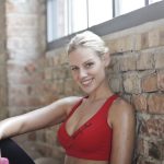 Blonde woman in red sportswear smiling with a dumbbell in a bright gym setting.
